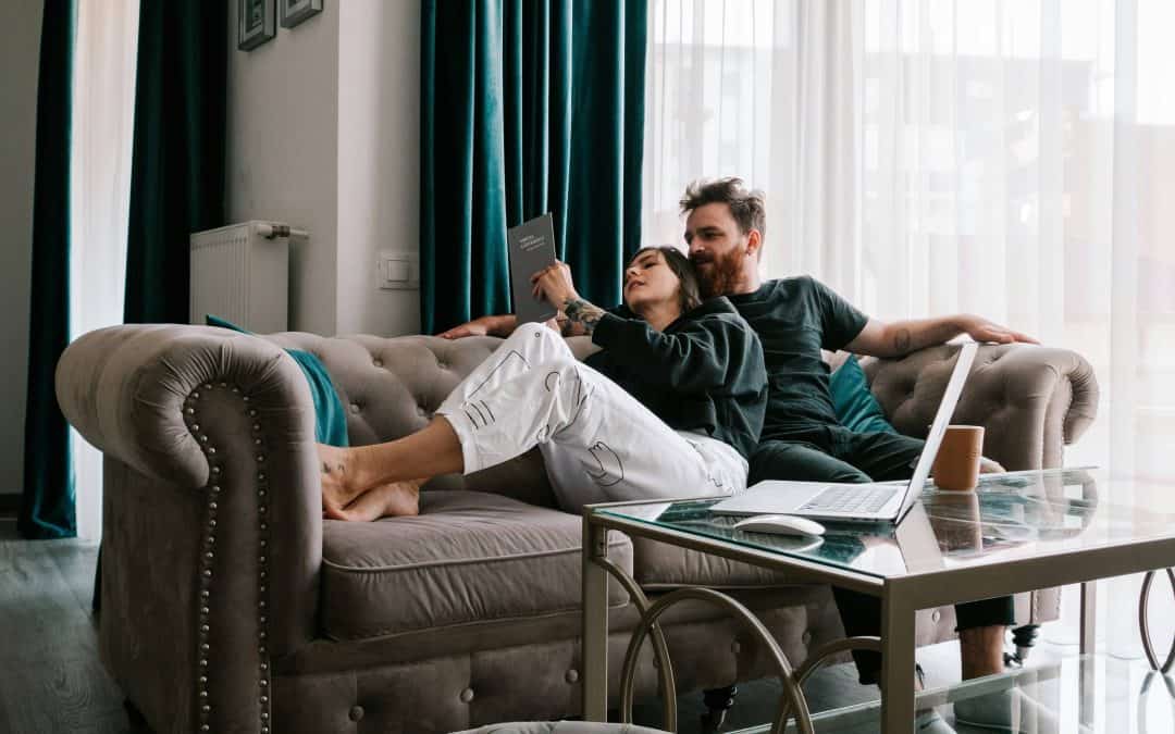 Cozy living room with couple relaxing on sofa, laptops, and natural light from large windows.