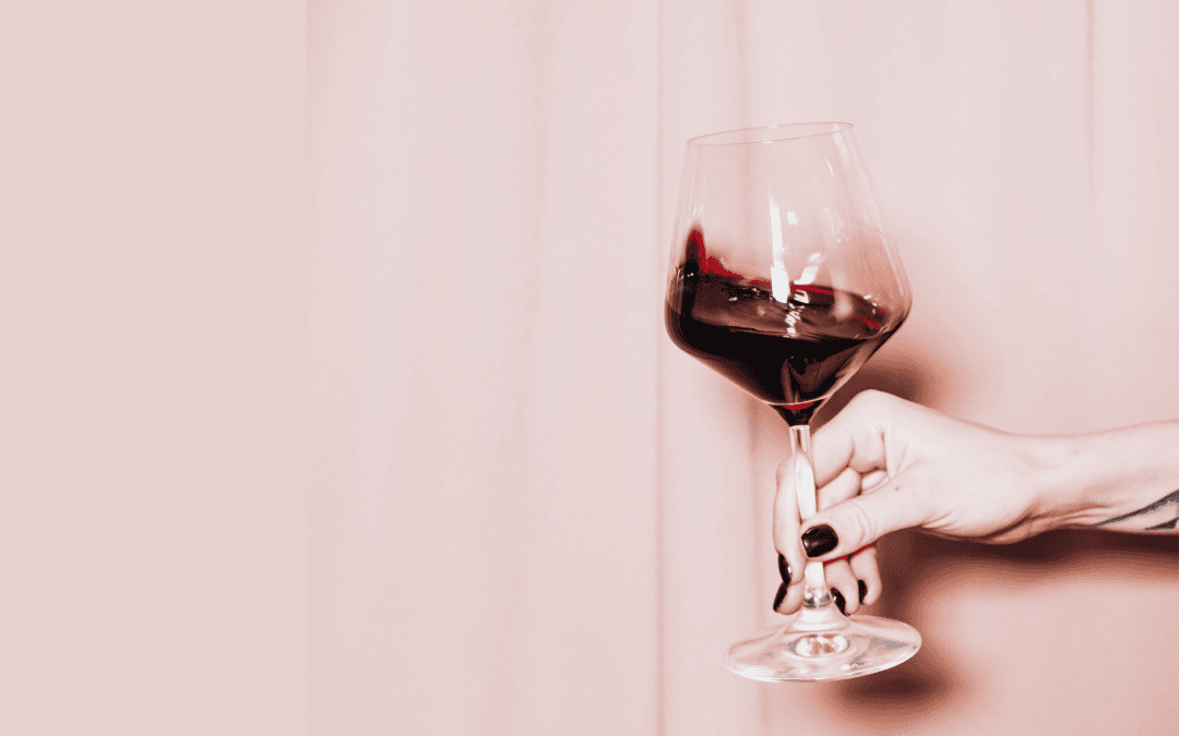 Rich red wine in a glass held by a hand with dark nail polish against a soft pink background.